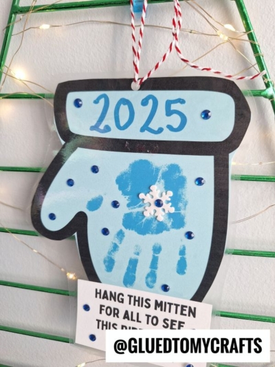 A handmade mitten height ornament labeled 2025 features a blue handprint, a white snowflake in the center, and blue gems. It hangs by red-and-white string with a note below and @GLUEDTOMYCRAFTS at the bottom.