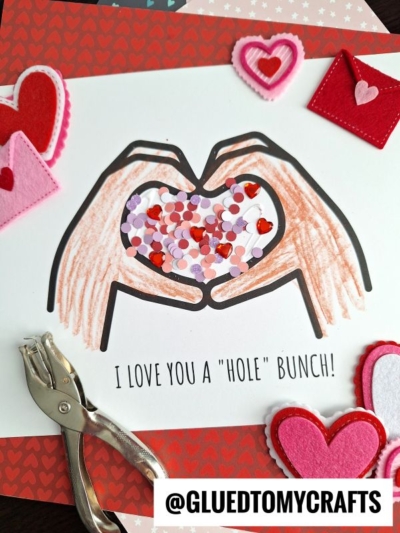 A handmade Valentines Day card shows two colored hands forming a heart shape, filled with heart-shaped confetti. Felt hearts, a hole punch, and decorative paper surround it. The text reads "love you a hole bunch!" for an extra-sweet touch.