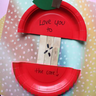 A paper plate craft resembling an apple core, with red edges and popsicle stick seeds. The top says Love you, and the bottom says to the core! on a colorful spotty background.