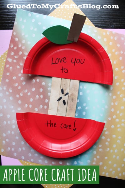 A paper plate craft resembling an apple core, with red edges and popsicle stick seeds. The top says Love you, and the bottom says to the core! on a colorful spotty background.