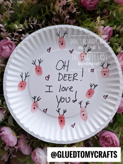 A white paper plate decorated with pink thumbprint reindeer, black marker faces, and antlers. In the center, it says OH DEER! I love you! Surrounded by pink flowers, this is one of the sweetest thumbprint craft ideas for spring.
