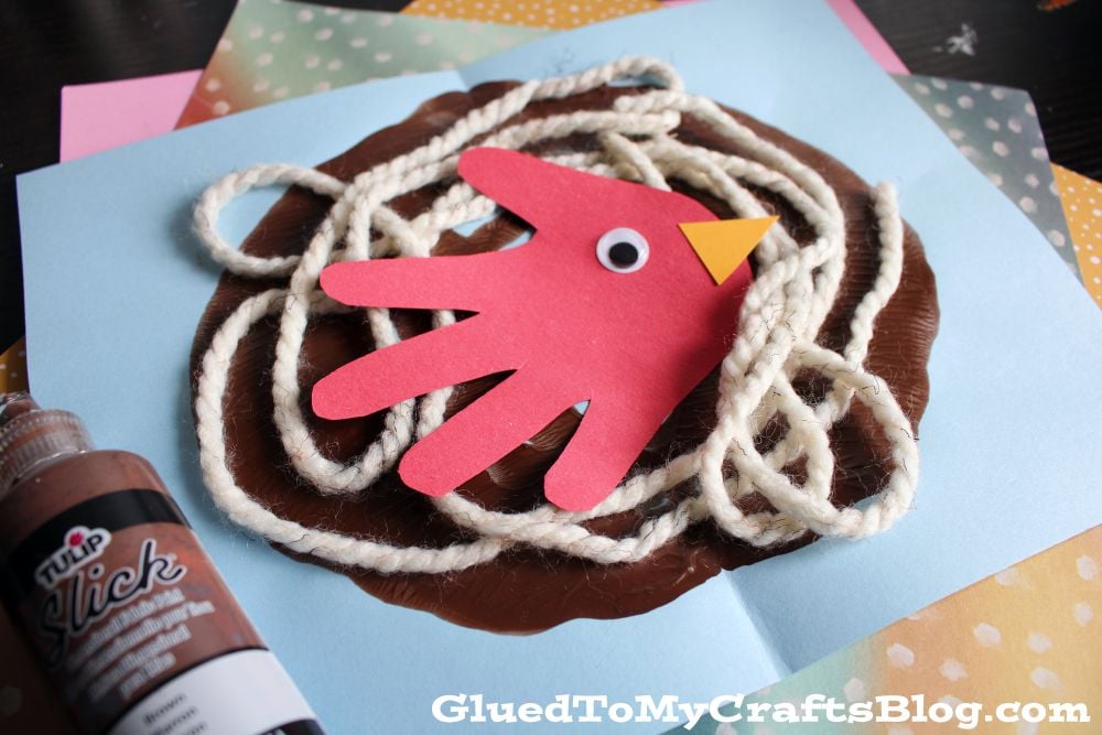 A red paper hand cutout with a googly eye and yellow beak, resembling a bird, sits atop wavy white yarn on brown paint, mimicking a nest, on colored paper with crafting supplies nearby.