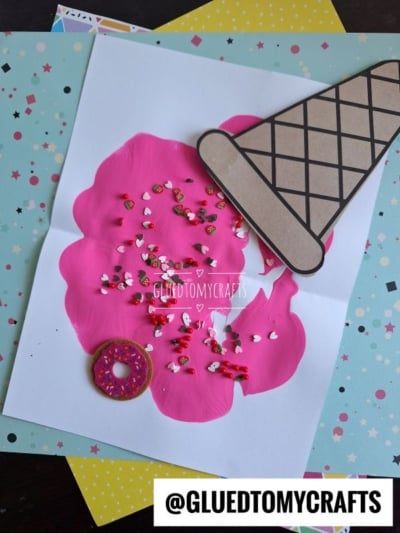 A handmade card features a large pink scoop of melted ice cream painted on white paper, topped with colorful sprinkles, a brown paper cone, and a small donut sticker. Decorative papers and playful paint splats are scattered underneath.