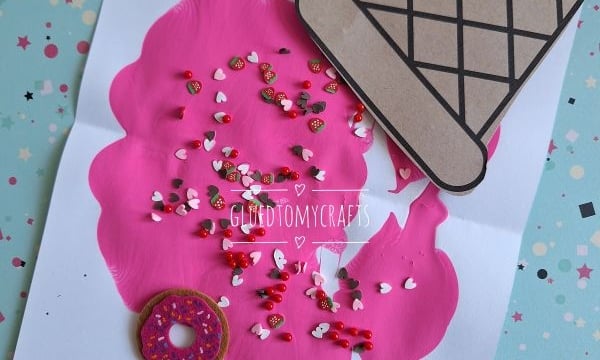 A handmade card features a large pink scoop of melted ice cream painted on white paper, topped with colorful sprinkles, a brown paper cone, and a small donut sticker. Decorative papers and playful paint splats are scattered underneath.