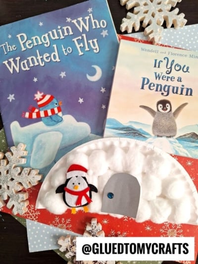 Two penguin-themed children’s books and a paper plate craft featuring a cotton ball igloo, penguin, and snowy igloo door are displayed on a festive snowflake background. A @GLUEDTOMYCRAFTS label is at the bottom.