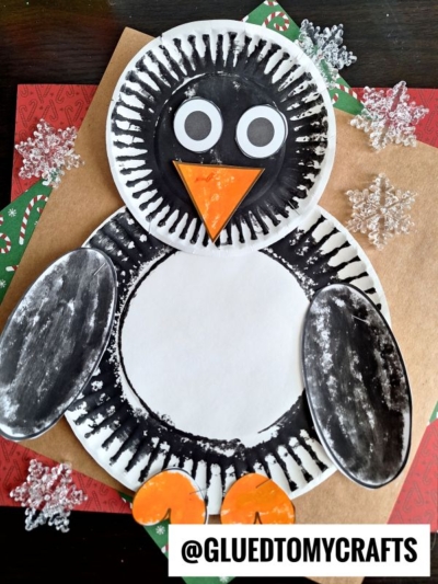 A charming paper plate penguin craft made with painted plates and orange paper for the beak and feet, surrounded by snowflake decorations and holiday-themed paper. The handle @GLUEDTOMYCRAFTS appears at the bottom.