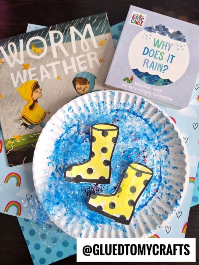 A paper plate rain puddle with blue textured art, two yellow rain boot cutouts, and two children’s books—“Worm Weather” and “Why Does It Rain?”—arranged on a rainbow-themed background. @GLUEDTOMYCRAFTS is labeled at the bottom.