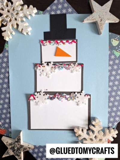 A paper snowman craft inspired by a snowman cake, made from three stacked white rectangles with a black hat, orange triangle nose, snowflake decorations, confetti details, and surrounded by star ornaments on a blue background.