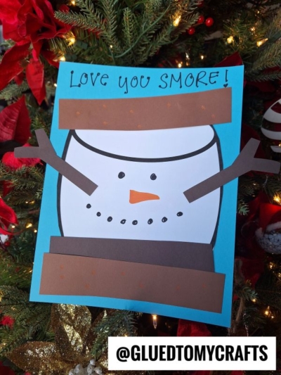 A paper snowman s'mores craft, with "Love you SMORE!" written on top, is displayed in front of a decorated Christmas tree. The @GLUEDTOMYCRAFTS logo appears at the bottom.
