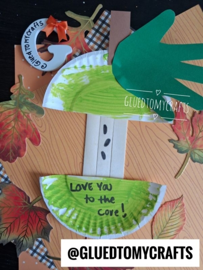 A fall-themed mother’s day apple core craft made with a paper plate, green handprint, leaves, a popsicle stick, and the words LOVE YOU TO THE CORE! surrounded by autumn leaves. @GLUETOMYCRAFTS is written at the bottom.