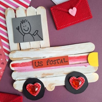 A handmade craft of a mail truck made from popsicle sticks, decorated with heart shapes for wheels and red envelopes, sits on a purple surface. A “US Postal” label and a drawing of a person are attached.
