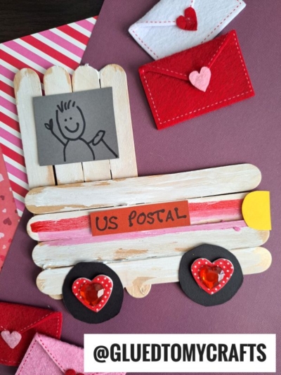 A handmade craft of a mail truck made from popsicle sticks, decorated with heart shapes for wheels and red envelopes, sits on a purple surface. A “US Postal” label and a drawing of a person are attached.