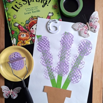 A spring-themed craft setup: a book titled Its Spring!, green tape, two decorative butterflies, a paper plate with purple paint, a potato masher, and a flower craft made with purple paint and a brown paper pot.