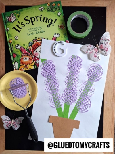 A spring-themed craft setup: a book titled Its Spring!, green tape, two decorative butterflies, a paper plate with purple paint, a potato masher, and a flower craft made with purple paint and a brown paper pot.