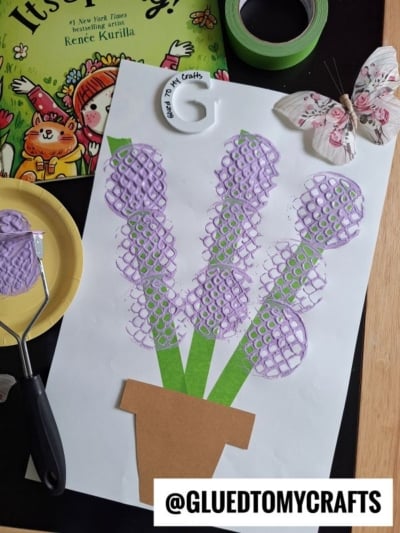 A childrens craft project shows purple flowers made by stamping paint with a mesh roller on white paper. The flowers have green stems and a brown paper pot. Craft supplies and a butterfly decoration surround the artwork.