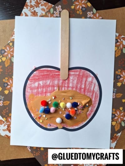 A puffy paint caramel apple paper craft with a popsicle stick, red crayon coloring, colorful pom-poms, and sprinkles as toppings. The image features floral-patterned paper and the text @GLUEDTOMYCRAFTS.