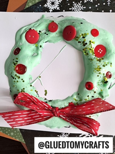 A handmade puffy paint holiday wreath, crafted from green puffy paint and adorned with red buttons, gold glitter, and a red ribbon bow at the bottom, is displayed on white paper with a snowflake pattern in the background.