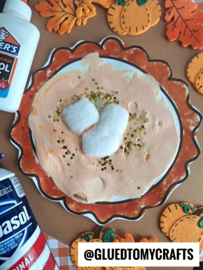 A puffy paint pie paper plate, painted like pumpkin pie and topped with cotton ball whipped cream and gold glitter, sits surrounded by felt pumpkin decorations, a bottle of glue, and a can of shaving cream.