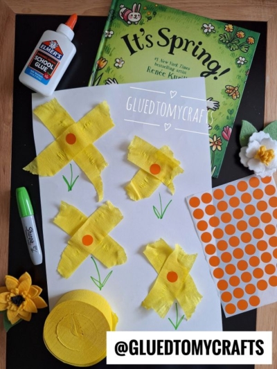 A spring craft setup with a book titled Its Spring!, yellow crepe paper craft ideas for flowers on white paper, green marker, orange dot stickers, glue, felt flower, and a @GLUEDTOMYCRAFTS label.