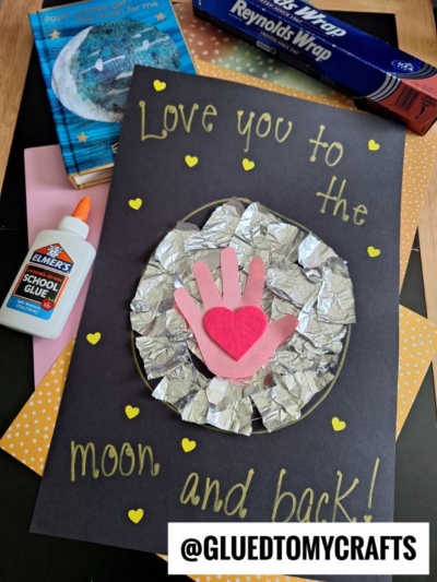 A handmade card with "Love you to the moon and back!" written in yellow, featuring an aluminum foil moon, pink paper handprints, a red heart, glue, colored paper, and foil in the background.