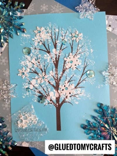 A winter tree craft on blue paper features a bare brown tree with white paint and snowflake decorations resembling snow, surrounded by plastic gems and snowflake embellishments. The handle @GLUEDTOMYCRAFTS is visible at the bottom.