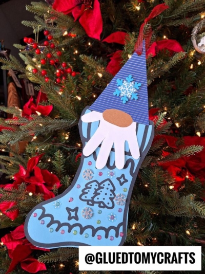 A blue gnome stocking ornament with a paper gnome face hangs on a decorated Christmas tree adorned with red flowers and lights. The handle is red and glittery. @GLUEDTOMYCRAFTS is written at the bottom.