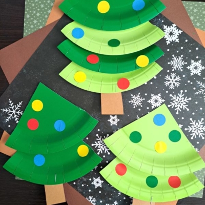 Three paper plate Christmas trees decorated with colorful dot stickers are displayed on snowflake-patterned and brown paper backgrounds. The trees have brown rectangular trunks. @GLUEDTOMYCRAFTS is written at the bottom.