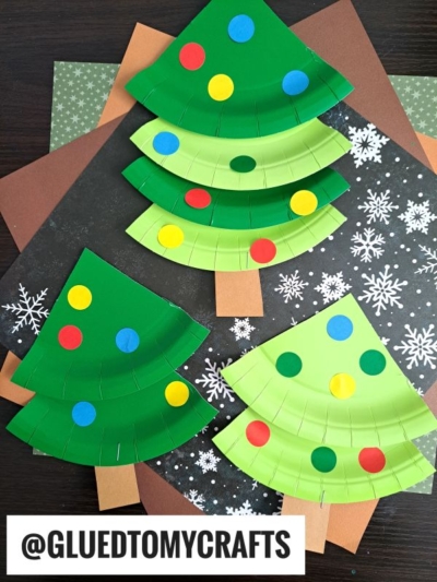 Three paper plate Christmas trees decorated with colorful dot stickers are displayed on snowflake-patterned and brown paper backgrounds. The trees have brown rectangular trunks. @GLUEDTOMYCRAFTS is written at the bottom.