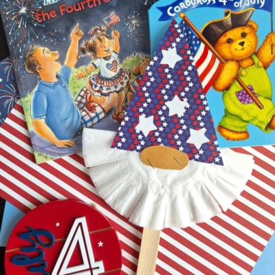 A festive flat lay featuring July 4th children’s books, a star-spangled gnome craft with a paper cup hat, striped paper, and a round “July 4” sign. The handle @GLUEDTOMYCRAFTS is visible.