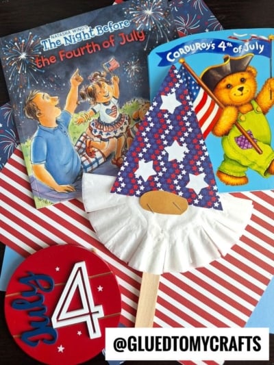 A festive flat lay featuring July 4th children’s books, a star-spangled gnome craft with a paper cup hat, striped paper, and a round “July 4” sign. The handle @GLUEDTOMYCRAFTS is visible.