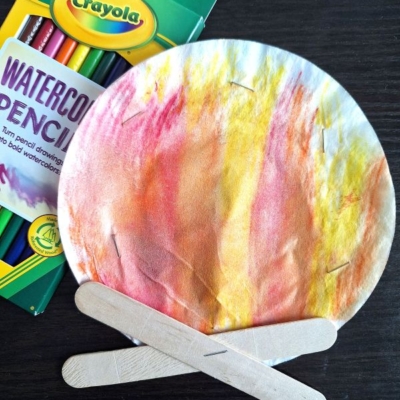 A paper plate painted with watercolor pencils to look like flames, with craft sticks arranged as logs, creating a campfire craft. A box of Crayola watercolor pencils is beside it. @GLUEDTOMYCRAFTS is at the bottom.