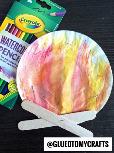 A paper plate painted with watercolor pencils to look like flames, with craft sticks arranged as logs, creating a campfire craft. A box of Crayola watercolor pencils is beside it. @GLUEDTOMYCRAFTS is at the bottom.