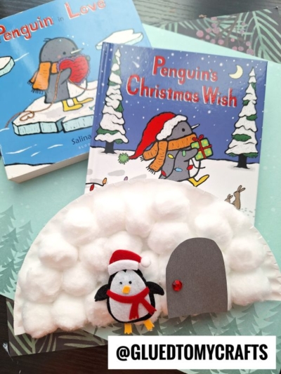 A charming cotton ball igloo craft, complete with a small felt penguin, is displayed in front of two children's books: Penguin in Love and Penguins Christmas Wish. @GLUEDTOMYCRAFTS.