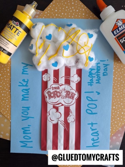 A handmade cotton ball Valentine’s Day card shaped like a popcorn box, with “Mom, you make my heart POP!” on blue paper, decorated with yellow paint, blue hearts, and “fresh popcorn” in the center. Craft supplies sit nearby.