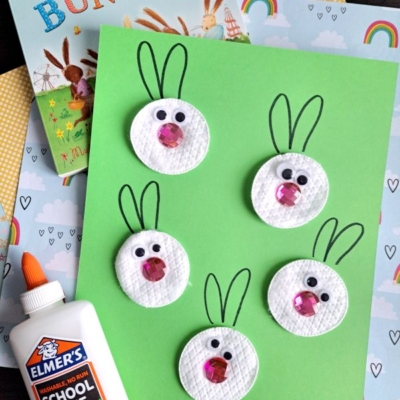 A green paper with five bunny faces made from white round materials, googly eyes, and pink noses. Drawn ears complete each face. Elmer’s glue, patterned papers, and a children’s Easter book are nearby.