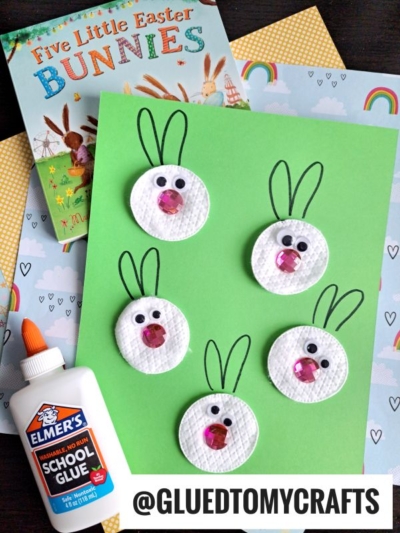 A green paper with five bunny faces made from white round materials, googly eyes, and pink noses. Drawn ears complete each face. Elmer’s glue, patterned papers, and a children’s Easter book are nearby.