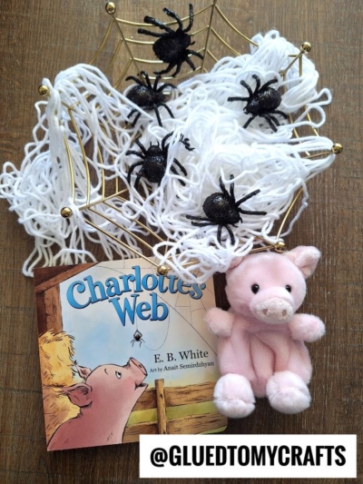 A copy of Charlotte’s Web, a small pink pig plush, white yarn arranged like a web, four black toy spiders, and a gold wire spider web are displayed on a wooden surface—perfect for story time sensory bin ideas. @GLUEDTOMYCRAFTS is at the bottom.