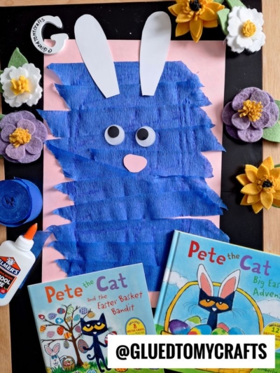 A blue Pete the Cat bunny crepe paper craft made from blue strips and white paper ears is surrounded by felt flowers, glue, blue yarn, and two Pete the Cat Easter books on a pink and black background.