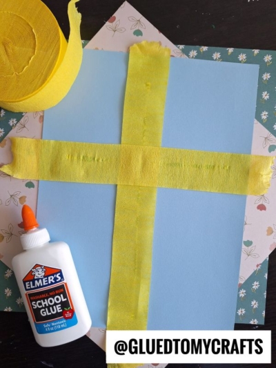 A piece of blue paper with yellow crepe paper taped in a cross shape, a bottle of Elmer’s school glue, floral-patterned paper underneath, and a roll of yellow crepe paper—perfect inspiration for crepe paper craft ideas. @GLUETOMYCRAFTS appears at the bottom.