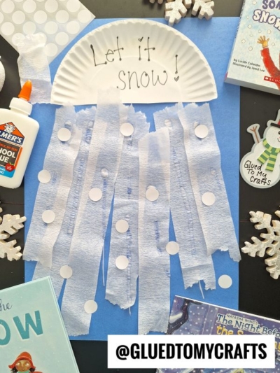 A paper plate with Let it snow! written on it is glued to blue paper. Strips of white tissue and paper circles hang down, resembling snow. Craft supplies and winter-themed books are around the artwork.