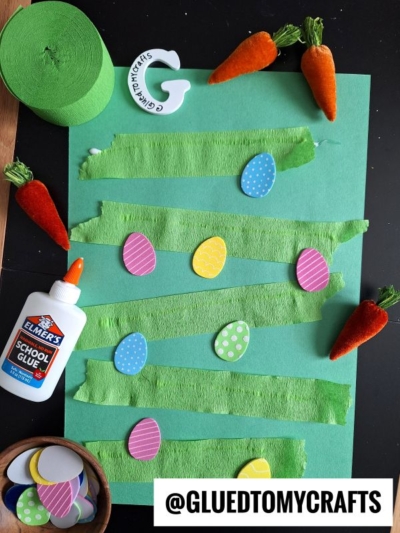 A green sheet of paper with strips of green crepe paper, colorful paper eggs, carrots, a bottle of Elmer's glue, foam circles, and an @GLUEDTOMYCRAFTS label arranged for creative spring crepe paper craft ideas.