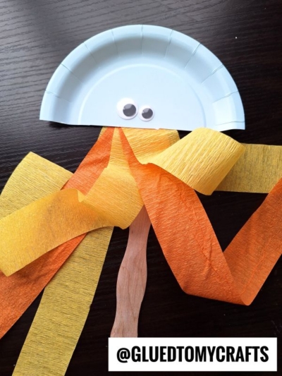 A light blue paper plate cut in half with two googly eyes, orange and yellow crepe paper streamers dangling below, and a craft stick handle—perfect for your crepe paper craft ideas. The text @GLUEDTOMYCRAFTS is at the bottom.