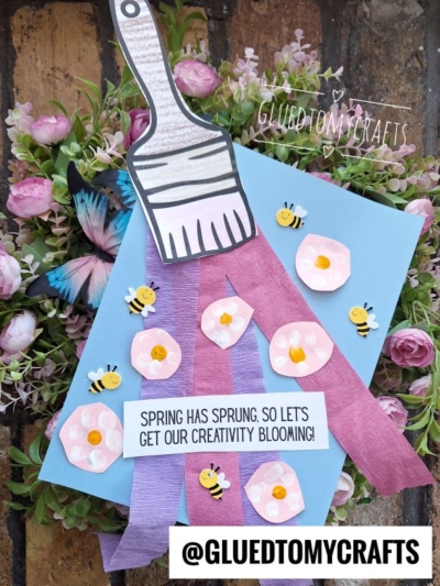 A spring-themed craft card with a paper paintbrush, purple paper strips, pink paper flowers with yellow centers, small paper bees, and a butterfly, surrounded by pink faux flowers. Text reads, Spring has sprung, so lets get our creativity blooming! and @GLUEDTOMYCRAFTS.