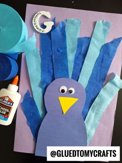 A paper peacock craft made with blue and light blue tissue paper strips, a purple body, yellow beak, and googly eyes on a purple sheet—perfect inspiration for crepe paper craft ideas. Includes glue, blue crepe paper rolls, and a paper letter G nearby.