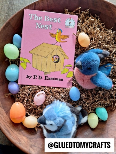 A bowl filled with colorful plastic eggs, brown shredded paper, two plush blue birds, and a pink book titled The Best Nest by P.D. Eastman—perfect for story time sensory bin ideas. The @GLUEDTOMYCRAFTS handle is at the bottom right.
