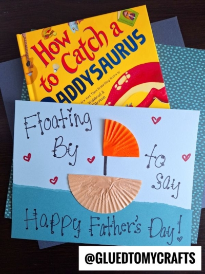 A handmade Fathers Day cupcake liner card craft with a blue background and a boat made from orange and beige cupcake liners, next to the book How to Catch a Daddysaurus. The card reads: Floating by to say Happy Fathers Day!.