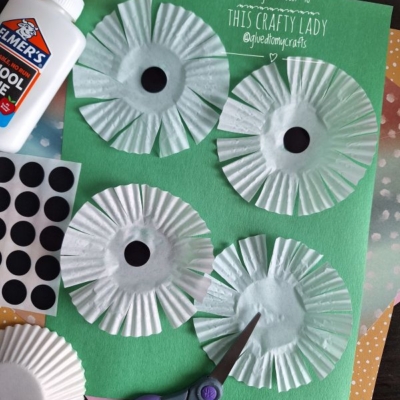 Four white cupcake liners cut into flower shapes are arranged on green paper, with glue, black dot stickers, scissors, and orange paper shown nearby as crafting materials.