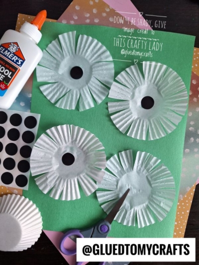 Four white cupcake liners cut into flower shapes are arranged on green paper, with glue, black dot stickers, scissors, and orange paper shown nearby as crafting materials.