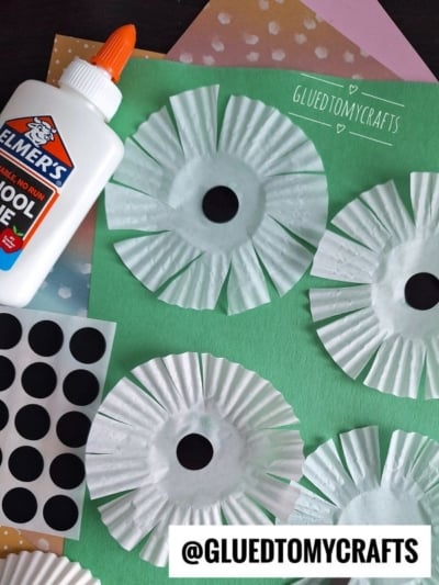 Several white paper flowers made from cupcake liners with black sticker centers are arranged on a green paper background. A bottle of Elmer’s glue and a sheet of black dot stickers are nearby.