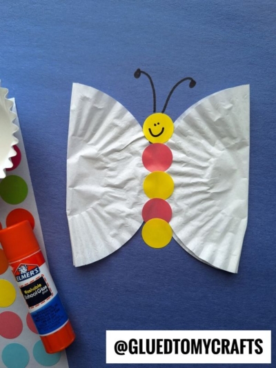 A butterfly craft made from white cupcake liners for wings and colorful paper circles for the body on a blue background. A glue stick and sticker sheet are beside it. The handle @GLUETOMYCRAFTS is at the bottom.